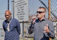 Markus Hilbert speaks into a microphone standing next to Mychal Johnson. Both stand in front of a chainlink fence with a sign that reads in part, "Flush truck debris from Con Edison underground structures, all other wastes not accepted."